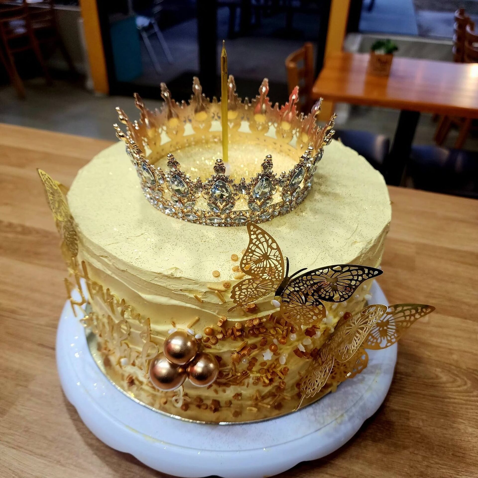 A 1Chick 1Bro gold crown 'Happy Birthday' cake with gold butterflies on a café table
